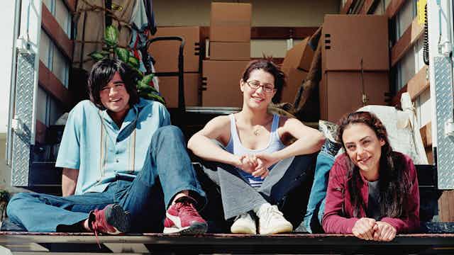 three young people sit in the back of a moving van with boxes in the background
