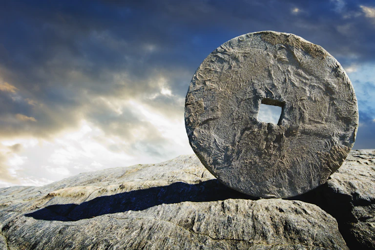 Against a blue and gray sky, an ancient wheel stands upright on a rocky patch of ground.