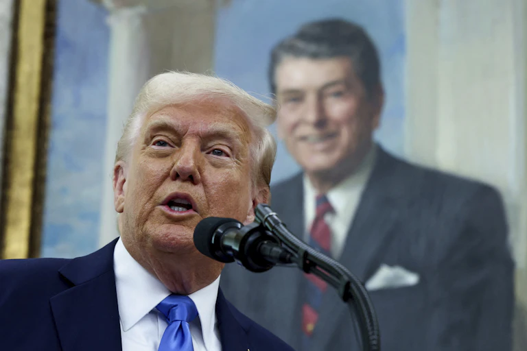 A man speaks into a microphone with a portrait of a man behind him.