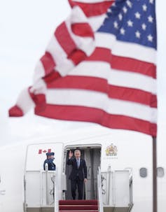 Un hombre con un corto cabello oscuro mientras el avión con una gran bandera estadounidense aterriza en primer plano.