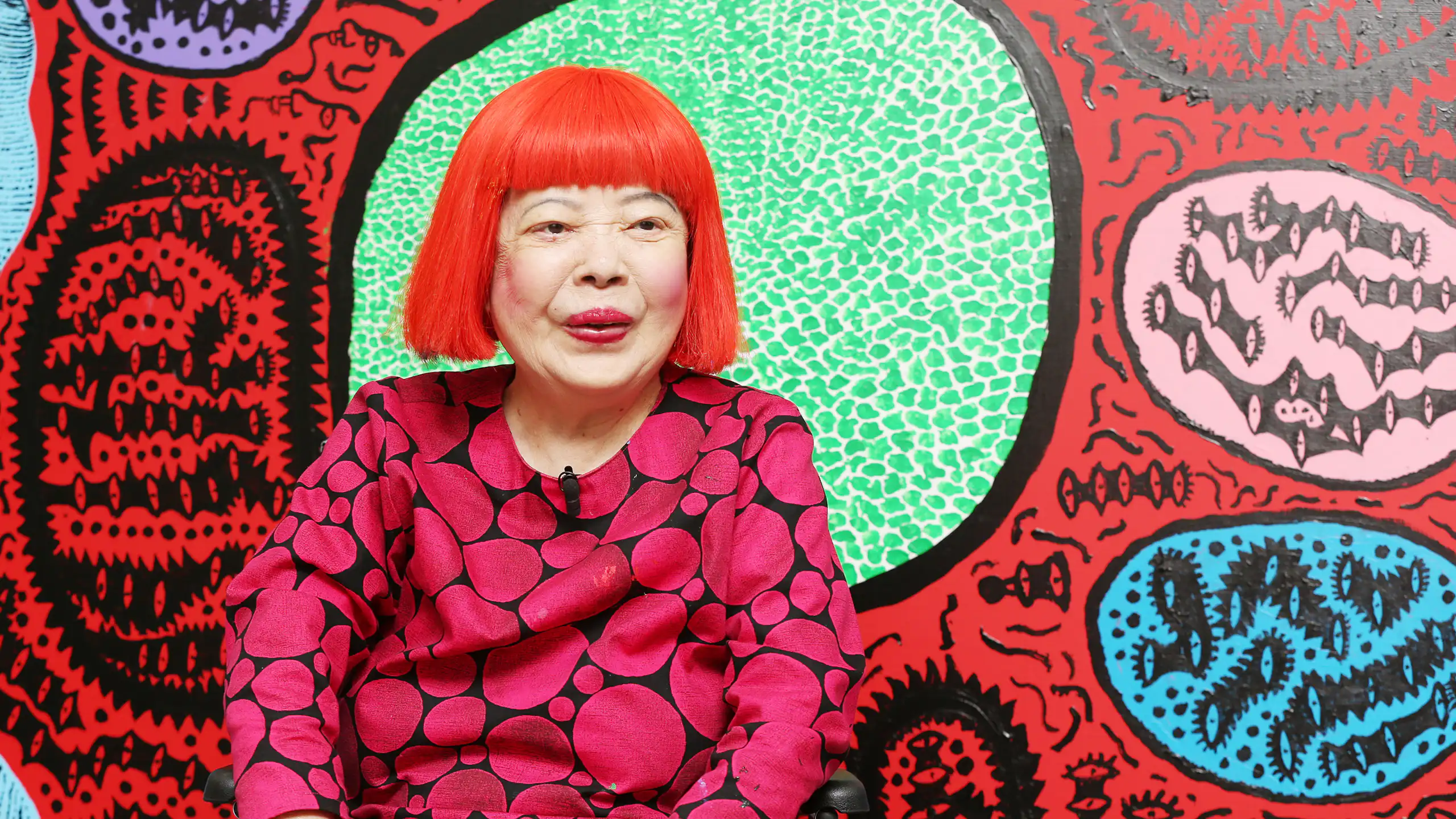 Kusama in a red wig in front of her paintings.