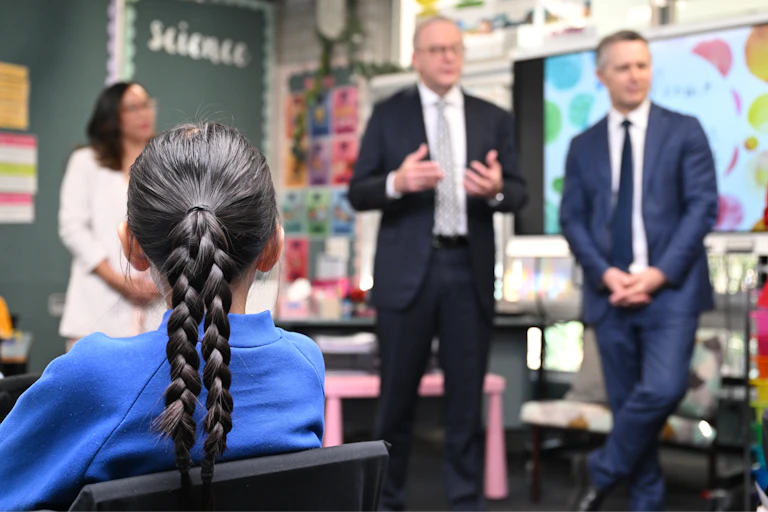 A young students listens to Prime Minister Anthony Albanese talking in her classroom. Education Minister Jason Clare stands next to Albanese.