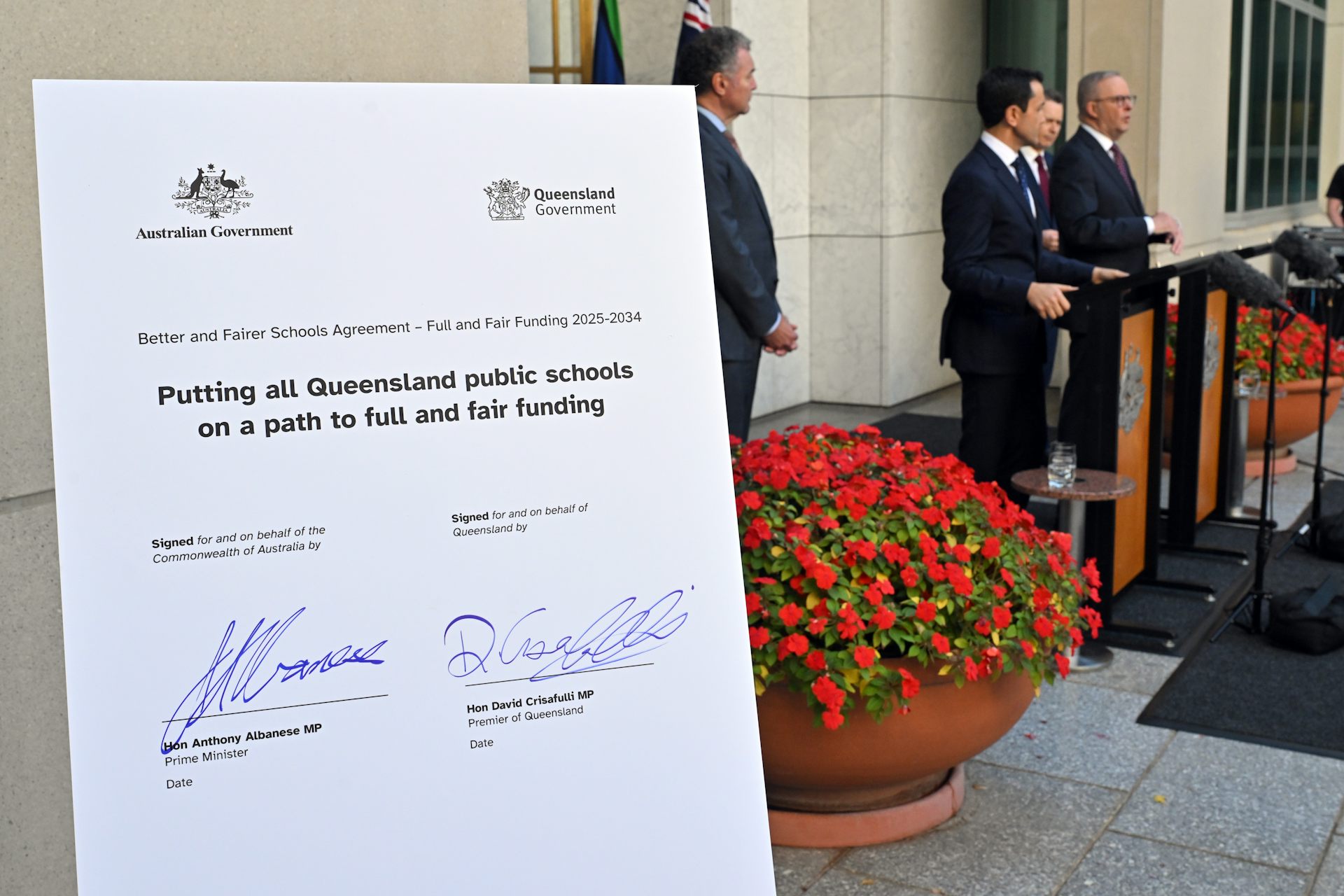 Prime Minister Anthony Albanese and Queensland Premier David Crisafulli speak at a press conference. A placard with their school funding agreement stands in the foreground.
