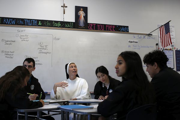 A woman in a white dress and black head covering sits at a table in a classroom with five teenagers seated around her.
