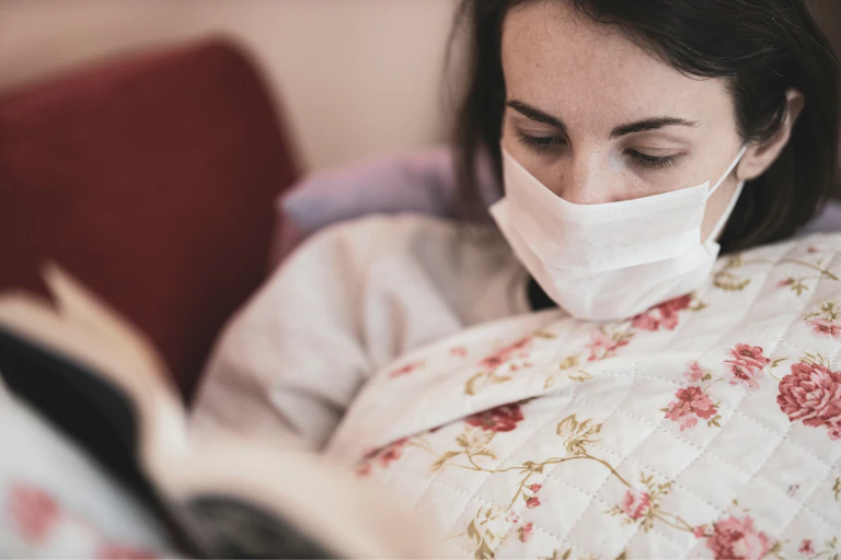Woman in a mask sits under a comforter, reading a book