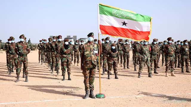 Armed men wearing face masks standing in formation, with one man at the front holding up a flag with green, white and red stripes and a black star in the middle