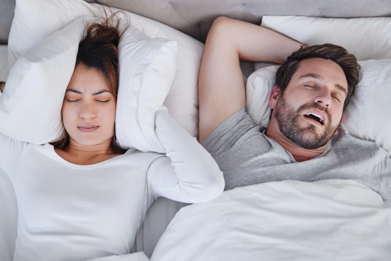 A couple in bed. The man is snoring and the woman covering her ears with a pillow.