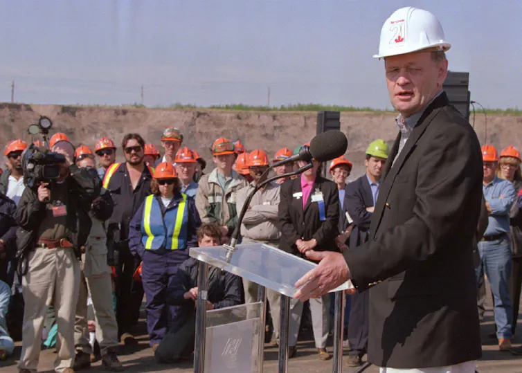 A man in a white hard hat stands at a podium talking to workers in orange hard hats.