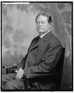 Un homme dans une veste de costume long, avec des cheveux noirs et des lunettes, pose pour une photo sur une chaise.