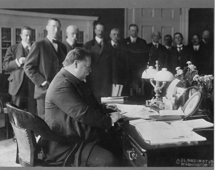 A large man at a paper-strewn desk, with many men standing near him.