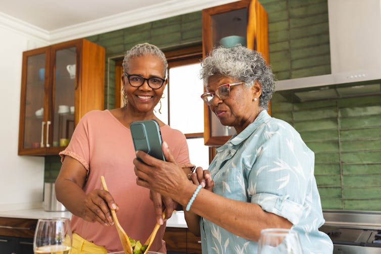 Two women in a kitchen, one holding serving utensils and the other showing her something on a smartphone