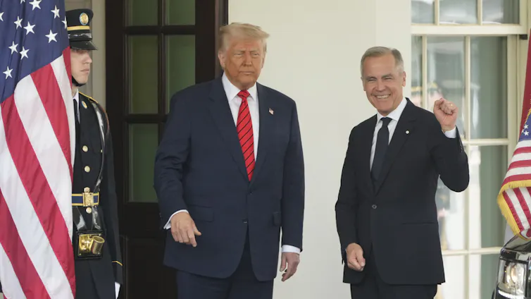 Two men stand smiling in the door way of a white stone building with American flags on either side of them. One man is corpulent with fluffy white-blond hair. The other has short grey hair and his fist is raised.