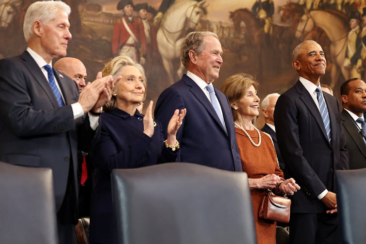 Three men in suits smile and clap as they stand next to two women also wearing formal clothing and are clapping and smiling.