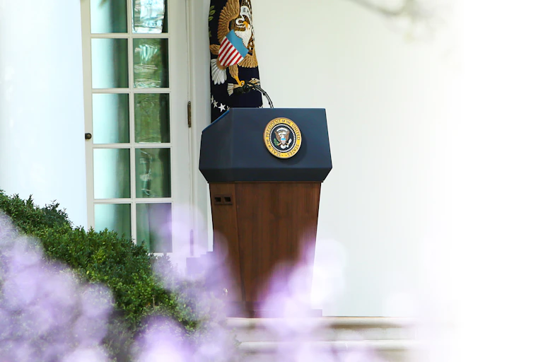 A podium shows the seal of the president of the United States and a microphone against a white wall and a blue flag.