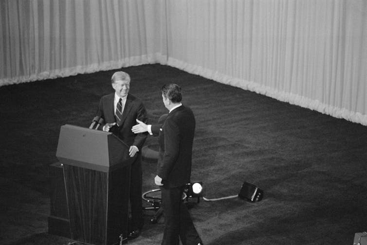 Two men in suits shake hands at a podium in a black-and-white photo.