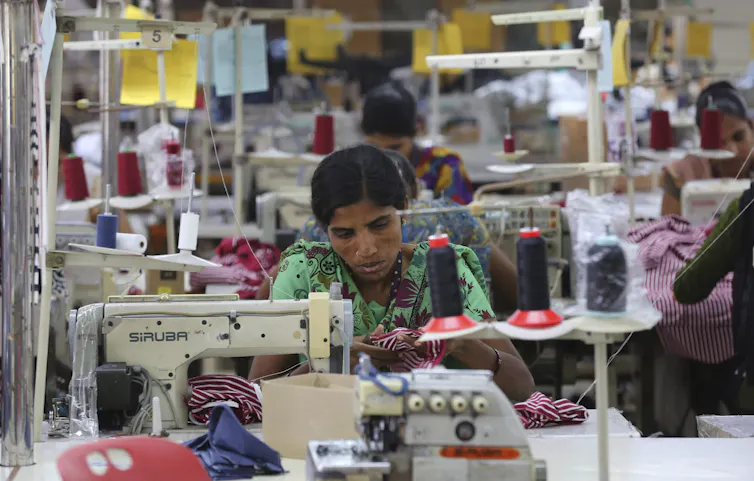 A woman operates a sewing machine in a factory surrounded by other workers