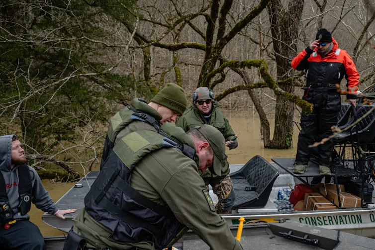a group of men wearing uniforms and life vests stand around boats in muddy water