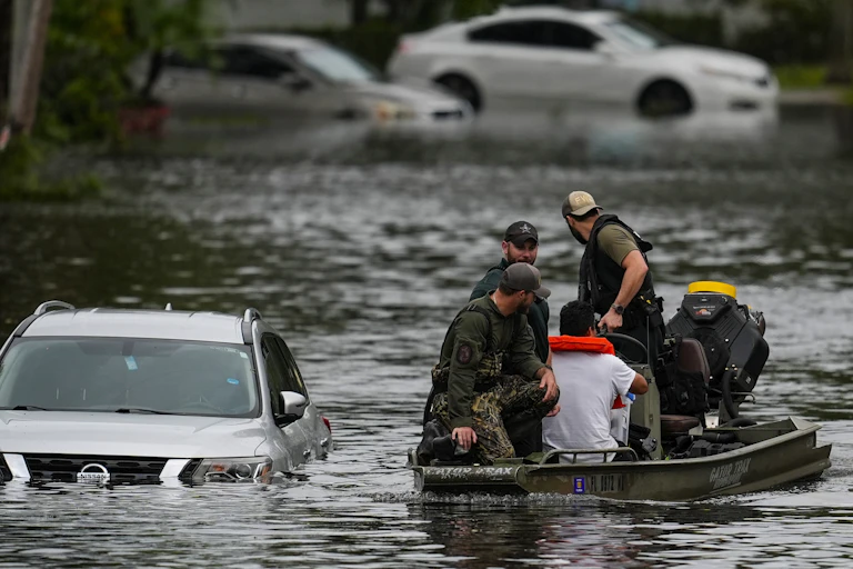 four men in a small boat traverse flood water by partially submerged a car