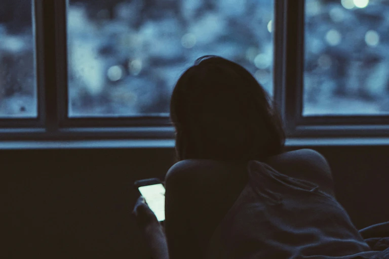 Woman by a window with dark city lights on her phone engaged in something.