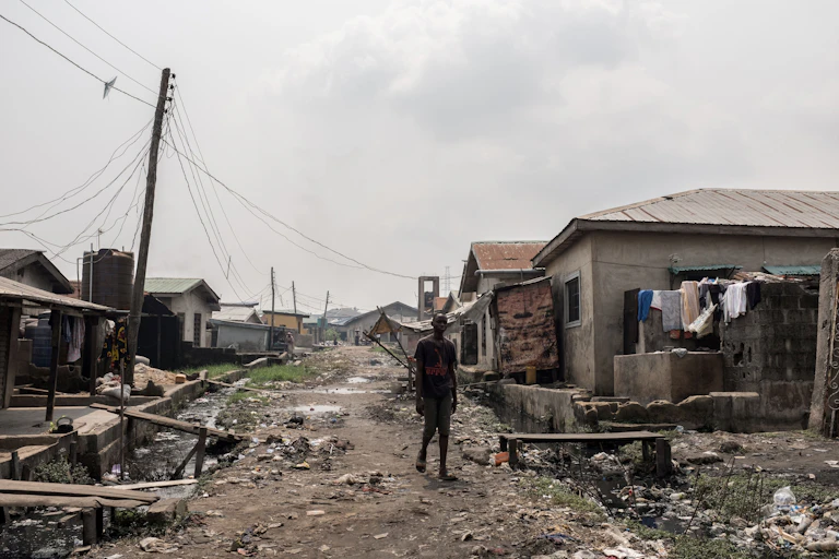 A man walks on a road between dilapidated buildings, with open drains and litter