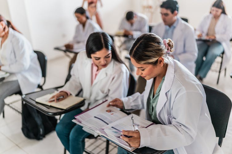 Medical students sitting in desks wearing white lab coats, discussing a textbook.
