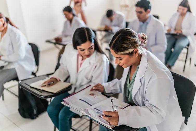 Medical students sitting in desks wearing white lab coats, discussing a textbook.