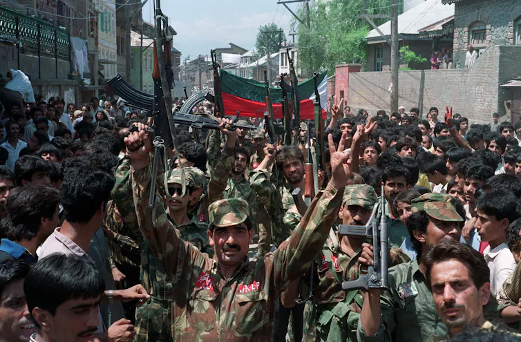 Men in army fatigues hold guns aloft while walking with people on a crowded street.