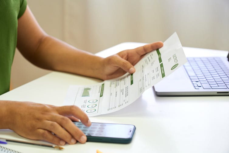 Torso of person in green shirt at table holding utility bill and phone with calculator app