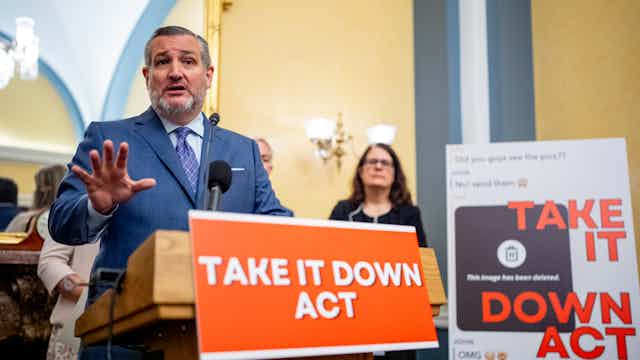 a man in a business suit speaks from a lectern as a man and a woman look on from behind him