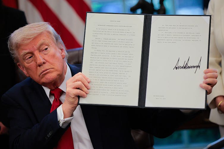 President Donald Trump wearing a blue suit and red tie displays a signed executive order.