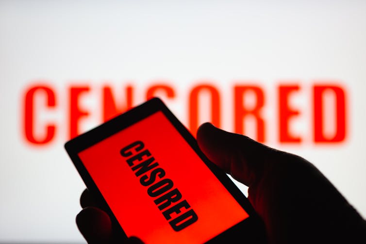 In the foreground, a hand hold a smartphone with the word 'censored' writtern on it, while the word 'censored' is seen on a white wall in the background.