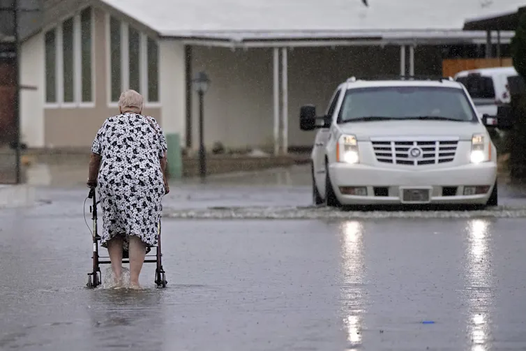 A woman using a walker walks through water up to her ankles. A car with lights on shows the water is deeper in nearby areas.