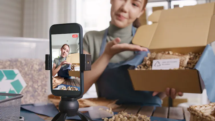 A smartphone on a stand records a woman opening a box and displaying the contents