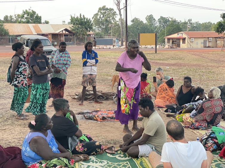 A woman speaking with a group of people, some seated and some standing, outdoors in a park or field.
