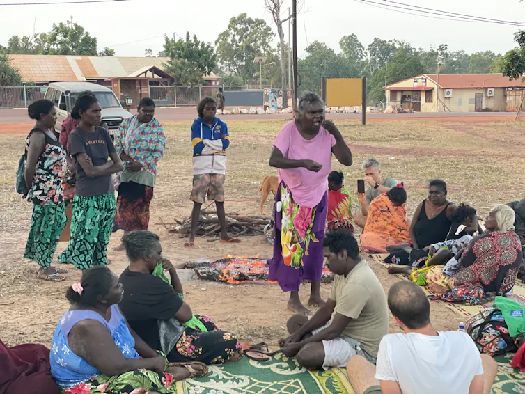 A woman speaking with a group of people, some seated and some standing, outdoors in a park or field.
