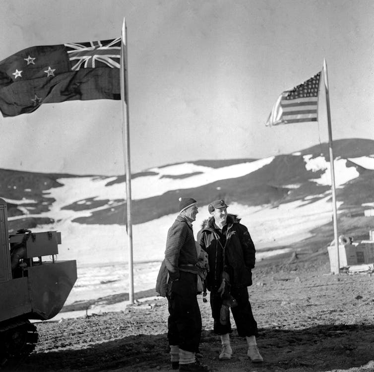 A black and white photo of two men standing between US and NZ flags.