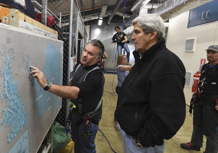A man in a dark jacket watches as another man points at a map.