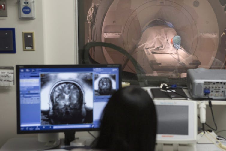 Máquina de resonancia magnética en segundo plano con una persona sentada en la pantalla que muestra la imagen del cerebro a la vanguardia