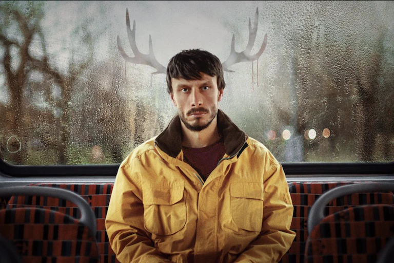 A man sitting at the back of a bus with a pair of antlers drawn on the window behind him.