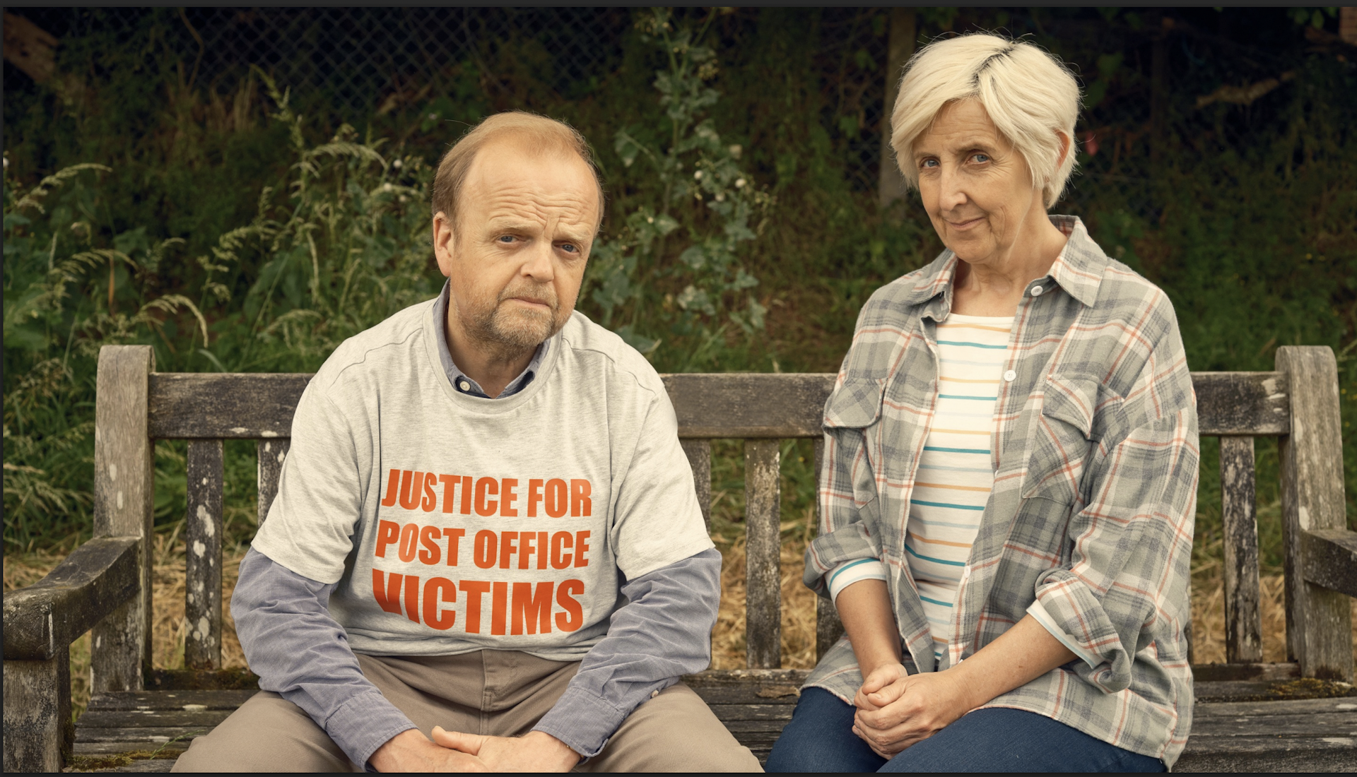 Two people looking pensive staring at the camera, one wearing a T-shirt saying 'Justice for the Post Office Victims'.