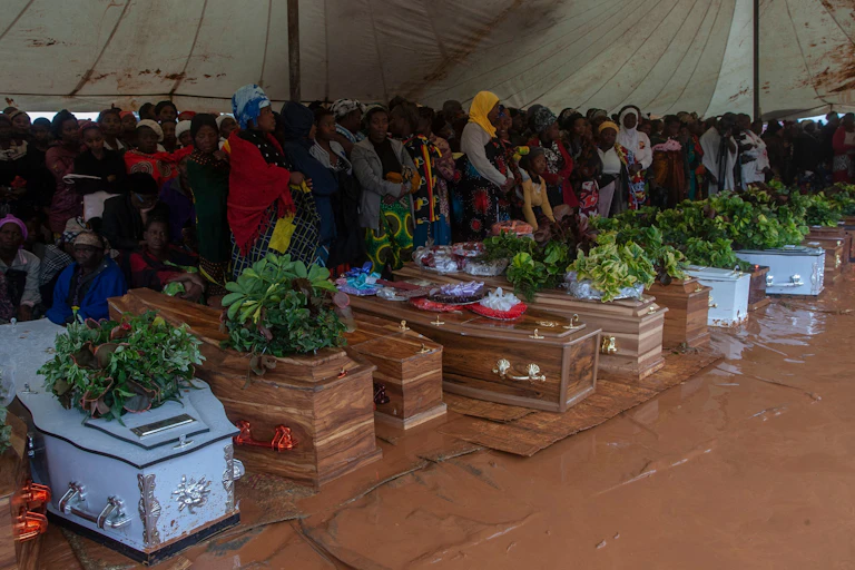 People sit in a marquee at a funeral of a row of 10 coffins which are on the ground in the mud