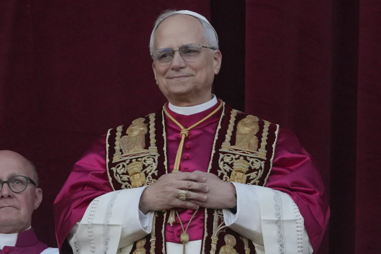 A man, dressed in brocaded vestments, stands with his arms folded and smiles gently.