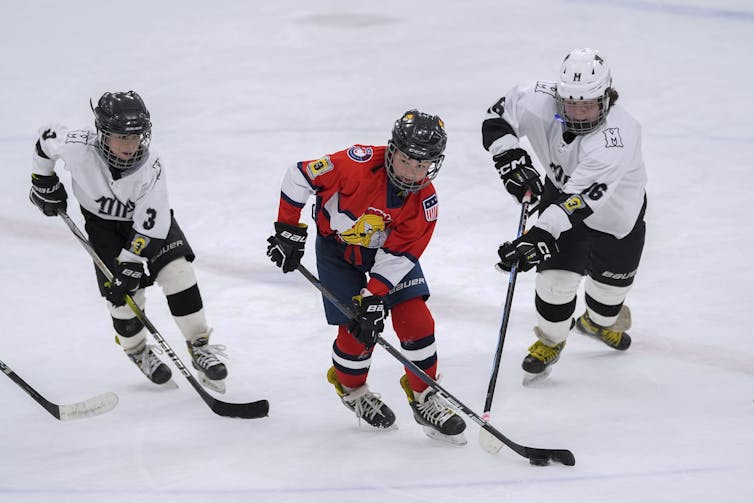 Niño en una pista de hielo única de hockey rojo, mientras que dos niños más con uniformes blancos se deslizan para ellos