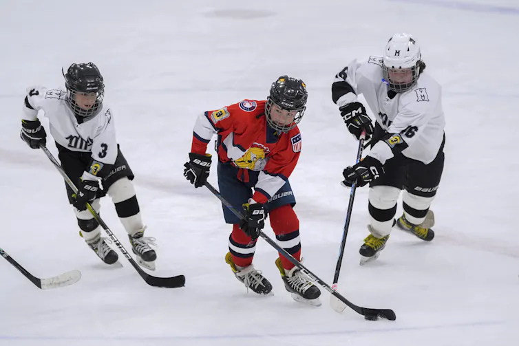 A child in a red hockey uniform skates with a puck while two other children in white uniforms skate after them