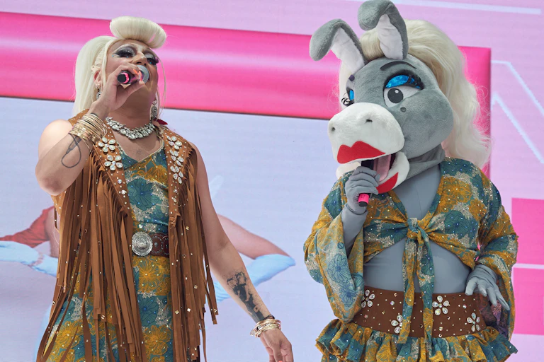 Two drag mascots -- one wearing a donkey costume -- appear on stage in front of a pink backdrop.