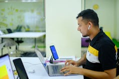 El joven está sentado en la mesa con vistas a una computadora portátil en auriculares en sus oídos.