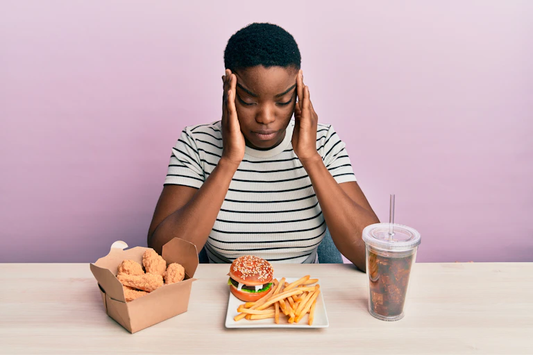 Young woman holds her temples as if she has a headache in front of a burger, fries and large coke