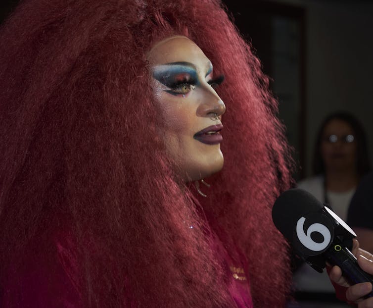 A drag performer, with bright red hair, speaks to an off-camera TV reporter.