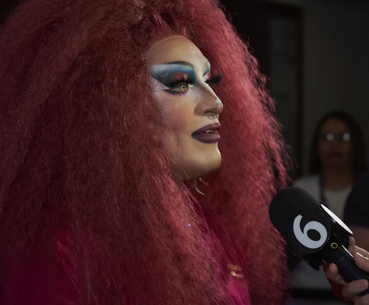 A drag performer, with bright red hair, speaks to an off-camera TV reporter.