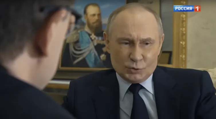 A screenshot of a man talking with a painting of a man iwth a beard behind him.